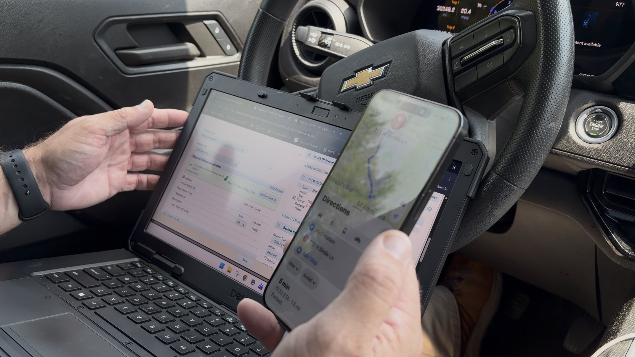 An inspector holding a phone with directions in front of a rugged in-truck laptop showing a permit record.